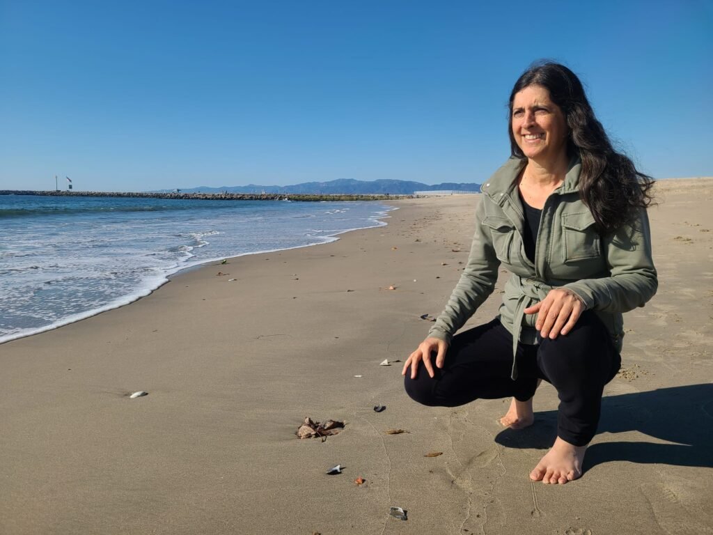 woman-with-long-brown-hair-kneels-on-a-beach-looking-forward